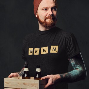 May include: A dark gray t-shirt with the word "WEEN" spelled out in Scrabble tiles. The person is holding a wooden crate with two brown beer bottles. The person is wearing a rust-colored beanie.