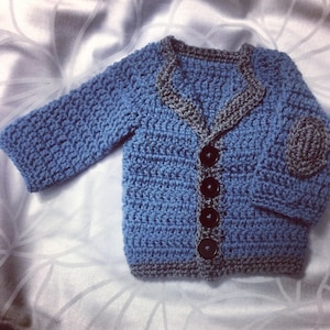 Puede incluir: Un cárdigan de punto azul con ribete gris y cuatro botones negros. El cárdigan tiene un parche en una manga.