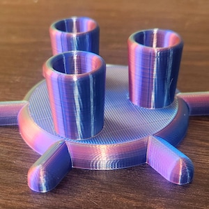 Könnte beinhalten: Ein dreizackiger, regenbogenfarbener Ständer aus Plastik in Blau und Rosa mit drei regenbogenfarbenen Bechern aus Plastik in Blau und Rosa.