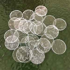 May include: A collection of clear glass circles of varying sizes, scattered on a green surface. The circles are translucent and have a slightly rippled texture.