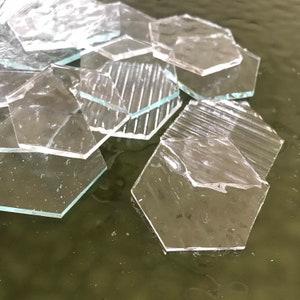 May include: A group of clear glass hexagonal tiles, some with a textured surface, are scattered on a green surface.