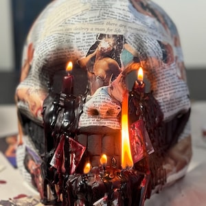 May include: A skull-shaped candle holder decorated with newspaper clippings and images. The skull has several lit candles with dark red wax dripping down. The text on the newspaper includes the phrase "because there are moments when".