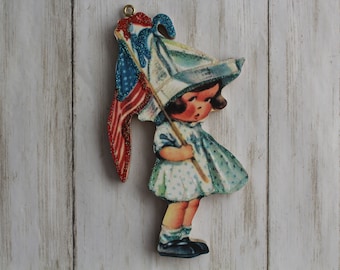 Puede incluir: Un adorno de madera de una niña que lleva un sombrero azul y blanco con una bandera estadounidense. La niña sostiene la bandera en su mano derecha y mira hacia abajo. El adorno está pintado en un estilo vintage.