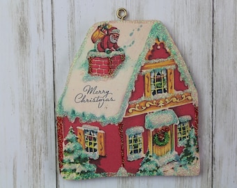 May include: A red and white Christmas ornament shaped like a house with a snow-covered roof. Santa Claus is climbing up the chimney. The ornament has glitter and the text "Merry Christmas" on the front.