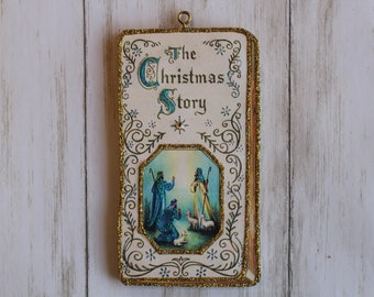 May include: A vintage Christmas ornament shaped like a book with a gold border and a white background. The book has the title "The Christmas Story" and a picture of three shepherds with a star in the sky.