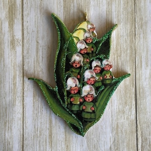 May include: A decorative ornament featuring a cluster of singing figures in green outfits and white bonnets, nestled within a green leaf-shaped design. The ornament has a gold-colored loop for hanging and is embellished with green glitter.