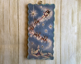 May include: A rectangular Christmas ornament featuring Santa Claus in a sleigh pulled by reindeer against a starry blue sky. The ornament has a scalloped edge with glitter and a gold-colored loop for hanging.