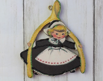 May include: A vintage-style Christmas ornament featuring a girl in a black and white dress holding a wishbone. The ornament has a gold glitter finish and a small gold loop for hanging. The girl's dress has a white apron with red cross-stitch detailing.