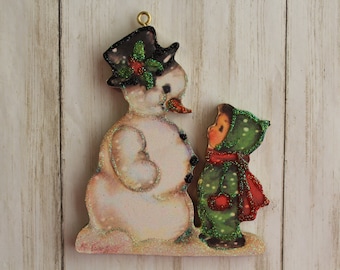 May include: A vintage-style Christmas ornament featuring a snowman with a black top hat and a green holly sprig, standing next to a child wearing a green hooded coat with red accents and white polka dots.