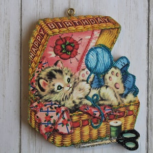 May include: A vintage-style Happy Birthday card featuring a kitten in a basket with yarn, scissors, and a spool of thread. The card is decorated with glitter and has a pink and green color scheme.