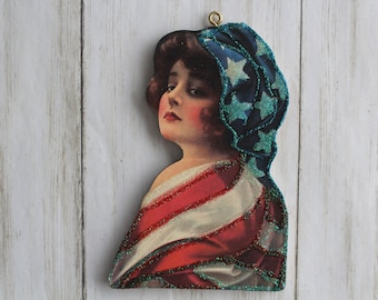 Puede incluir: Un adorno de madera de estilo vintage con el rostro de una mujer con un pañuelo de cabeza azul y blanco con estampado de estrellas. La mujer lleva un vestido rojo, blanco y azul con detalles de purpurina.