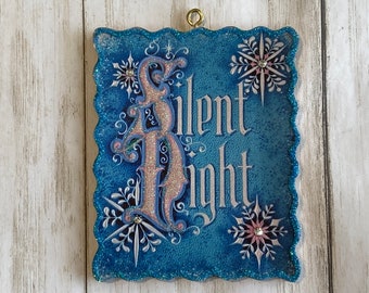 May include: A blue Christmas ornament with the words "Silent Night" in silver script. The ornament has a glittery border and features snowflakes with rhinestone accents. A gold loop is at the top for hanging.