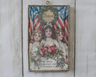 Puede incluir: Una impresión enmarcada de estilo vintage con un borde dorado brillante. Tres chicas sostienen un ramo de flores rojas y blancas. La impresión presenta una corona con la palabra "Paz" y dos banderas americanas. El texto en la parte inferior dice "from every mountain side let freedom ring!"