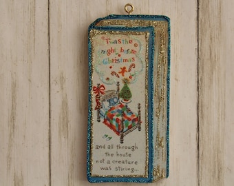 May include: A vintage Christmas ornament with a blue and gold frame. The ornament features a picture of a child sleeping in a bed with a Christmas tree in the background. The text on the ornament reads "Twas the night before Christmas and all through the house not a creature was stirring..."