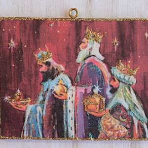 May include: A vintage Christmas ornament featuring a red background with three wise men in colorful robes and gold crowns. The men are carrying gifts and walking towards a star in the sky.