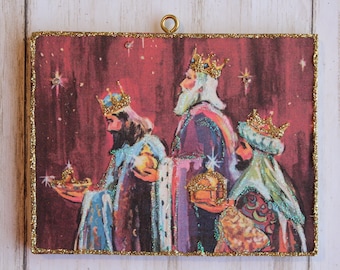 May include: A vintage Christmas ornament featuring a red background with three wise men in colorful robes and gold crowns. The men are carrying gifts and walking towards a star in the sky.