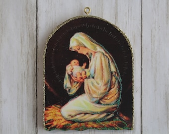 May include: A vintage-style Christmas ornament featuring a depiction of Mary holding baby Jesus. The ornament is made of wood and has a gold glitter border. The image is set against a dark background and features a halo around Mary's head.
