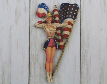 May include: A wooden ornament featuring a woman in a red, white, and blue uniform holding an American flag. The ornament is decorated with glitter and has a gold string for hanging.