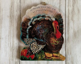 May include: Thanksgiving-themed wooden decoration featuring a colorful turkey with a red wattle and a fan of feathers. The decoration includes the text "ACROSS THE MILES at Thanksgiving". A pumpkin and foliage are at the base.