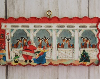 May include: A rectangular Christmas ornament with a red scalloped border and gold hanging loop. The ornament depicts Santa Claus with a sack of gifts, reindeer, and the names of the reindeer: Dasher, Dancer, Prancer, Vixen, Comet, Cupid, Donner, and Blitzen.