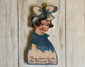 May include: A vintage paper doll ornament with a blue dress and a white bow. The doll has a blue glitter finish and the text "Three cheers for the Red, White and Blue." is printed on the bottom.