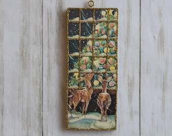 May include: A vintage Christmas ornament featuring a window scene with two deer looking at a decorated Christmas tree. The window is framed with gold glitter and the scene is set against a snowy background.
