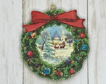 May include: A green glitter Christmas wreath ornament with a red bow and a winter scene inside. The scene depicts a snowy village with a church and a horse-drawn sleigh.