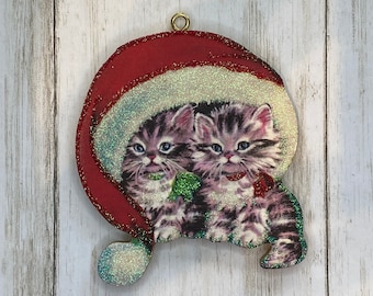 May include: A Christmas ornament featuring two kittens wearing a Santa hat. The ornament has a red and glittery white trim. The kittens are gray and white with blue eyes and green and red bows. The ornament has a gold loop for hanging.