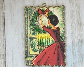 May include: A vintage Christmas ornament featuring a woman in a red dress hanging a wreath on a door. The ornament is decorated with glitter and has a gold border.