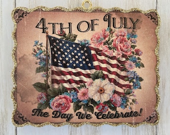 Può includere: Un cartello in stile vintage per il 4 luglio con una bandiera americana rossa, bianca e blu circondata da fiori rosa, bianchi e blu. Il cartello recita "4th of July The Day We Celebrate!"