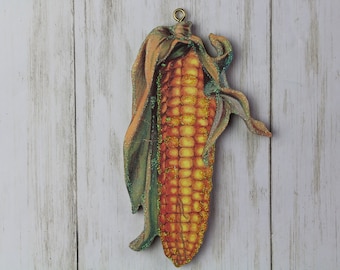 May include: A wooden corn decoration with a gold-colored loop for hanging. The corn has a yellow and orange color scheme with green and gold accents. The husk is a light brown color. The background is a white wooden surface.
