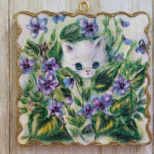 May include: A white kitten with blue eyes peeks out from behind a cluster of purple and green flowers. The image is surrounded by a gold glitter border.