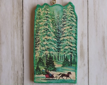 May include: A wooden Christmas ornament with a green glitter border. The ornament features a vintage illustration of a horse-drawn sleigh traveling through a snowy forest.
