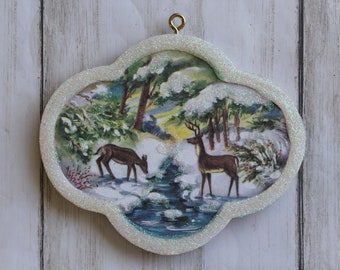 Könnte beinhalten: Ornament mit einer Winterlandschaft, die zwei Hirsche in der Nähe eines Baches in einem verschneiten Wald zeigt. Das Ornament hat einen glitzernden weißen Rand und eine goldene Schlaufe zum Aufhängen. Eine festliche Weihnachtsdekoration.
