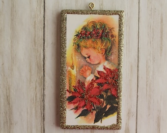 May include: A vintage Christmas ornament featuring a young girl with a red flower crown, praying with her hands clasped in front of a lit candle. The ornament is framed with a gold glitter border and features red poinsettia flowers.