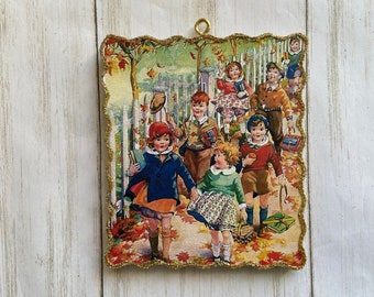 May include: Vintage-style decorative ornament featuring a charming illustration of children walking to school in autumn. The image has a gold glitter border and a small gold loop for hanging. The children are dressed in vintage clothing and carrying books.