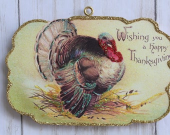 May include: Thanksgiving decoration featuring a turkey illustration with glitter accents. The image has a gold glitter border and the text "Wishing you a Happy Thanksgiving". Includes a gold loop for hanging.