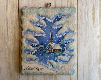 May include: A vintage Christmas ornament featuring a snowy church scene with a blue sky and white snow-covered trees. The ornament is made of glitter and has a scalloped edge. The text "Silent Night" is written on the bottom of the ornament.