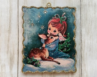 May include: A vintage Christmas ornament featuring a girl with red hair and a blue dress holding a fawn. The ornament has a glittery blue background with gold stars and a gold border.
