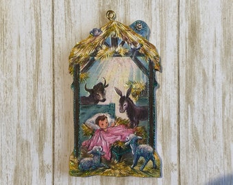 May include: A vintage Christmas ornament depicting the nativity scene. A baby Jesus lies in a manger with a cow, donkey, and two sheep surrounding him. The ornament is made of cardboard and has a glitter finish.