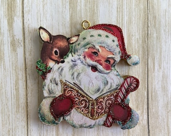May include: A vintage Christmas ornament featuring a red-suited Santa Claus reading a book with a small deer peeking over his shoulder. The ornament is decorated with glitter and has a gold hanger.