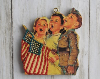 Puede incluir: Un adorno navideño de madera con una ilustración vintage de tres niños cantando con una bandera estadounidense brillante. Los niños visten ropa patriótica, incluido un uniforme militar.