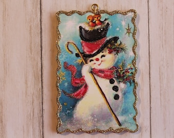 May include: A vintage Christmas ornament featuring a snowman wearing a red and black hat, a red scarf, and holding a stick. The ornament has a gold glitter border and a blue background with white snowflakes.