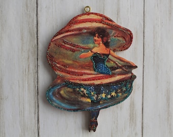 Puede incluir: Un adorno de madera con una ilustración vintage de una mujer con un vestido azul y una falda roja, blanca y azul. La falda está decorada con estrellas y purpurina.