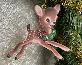 Könnte beinhalten: Ein hölzerner Weihnachtsbaumschmuck in Form eines Rehs mit einem grünen Glitzerkranz um den Hals. Das Reh hat braunes Fell mit weißen Flecken und eine rote Schleife am Kranz.
