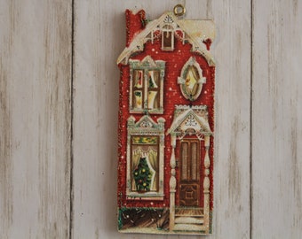 May include: A red and white Christmas ornament shaped like a house with a small Christmas tree in the window. The ornament has a glittery finish and a gold string for hanging.