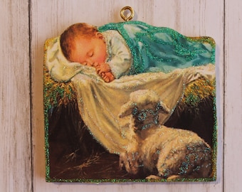 May include: A Christmas ornament depicting a sleeping baby Jesus in a manger with a white lamb beside him. The ornament is made of wood and has a glittery finish.