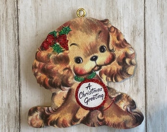 May include: A vintage-style Christmas ornament featuring a cartoon dog with brown fur and a red nose. The dog wears a green collar and a holly berry bow. The ornament has the text "A Christmas Greeting" on a red glitter circle.