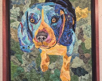 Pode incluir: Uma peça de arte de parede em colagem de tecido apresentando um retrato de um cão beagle em tons de azul, amarelo e laranja, sobre um fundo verde texturizado. A obra está inserida numa moldura retangular de madeira com acabamento castanho escuro.