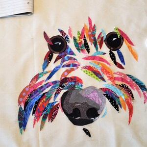 Pode incluir: Uma obra de arte de tecido colorida que representa o rosto de um c&atilde;o. A pelagem do c&atilde;o &eacute; feita de v&aacute;rios peda&ccedil;os de tecido coloridos, e os olhos s&atilde;o pretos com pupilas brancas. O nariz &eacute; um tecido cinza escuro com um padr&atilde;o floral.